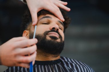 Erkek müşteri, modern bir berber dükkanında berberlik yaparken ustalıkla sakalını kesiyor ve şekillendiriyor.