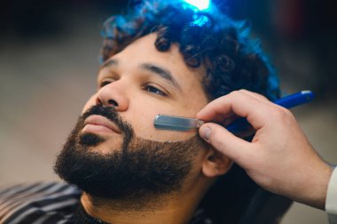 Berber, müşterinin sakalını ustura ile dikkatlice şekillendiriyor. Keskin çizgilere ve profesyonel bakım hizmetlerine odaklanıyor.