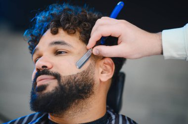 Erkek müşteri profesyonel sakal kesimi yaptırıyor ve berber dükkanında berberden ustura alıyor.