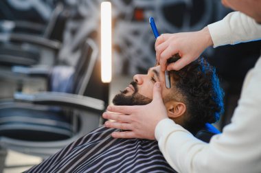 Modern bir berber dükkanında geleneksel bir ustura ile müşterinin sakalını dikkatlice tıraş eden yetenekli bir berber.