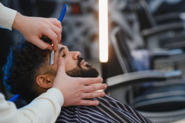 Berber, modern bir berber dükkanında bir adamın sakalını ve saçını ustura ile dikkatlice şekillendirir ve tıraş eder.