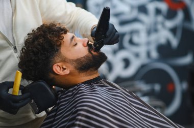 Berber dükkanı uzmanı, bir müşterinin sakalını elektrikli makasla dikkatlice buduyor ve profesyonel tımar hizmeti veriyor.