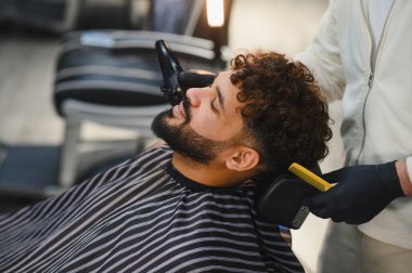 Berber dükkanı stilisti müşterinin kıvırcık saçlarını buduyor ve tarıyor, sakalını şekillendiriyor, profesyonel bakım hizmetleri sunuyor.