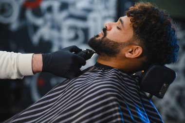 Berber koltuğunda oturan bir adam eldiven takan profesyonel bir berberden sakal tıraşı oluyor.