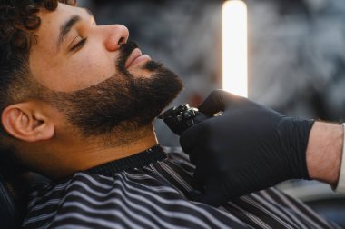 Modern bir kuaförde kırpıcı kullanarak erkek müşterinin sakalını şekillendiren ve şekillendiren profesyonel berber.
