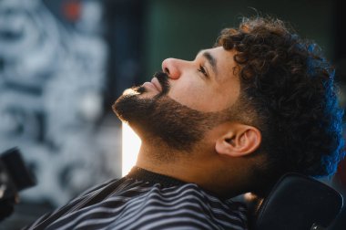 Salon sandalyesinde oturan genç adam sakalını kesiyor ve modern saç kesimi yaptırıyor. Rahat bir müşteri şık erkek bakımı ve kendine güveni gösteriyor.