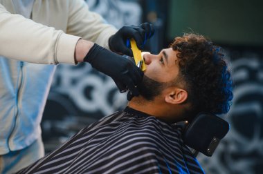 Berber, modern bir berber dükkanında ustaca tıraş ediyor ve bakımlı bir adamın sakalını tıraş ediyor. Yakın plan, profesyonel bakım.