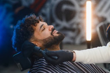 Saçları kıvırcık olan bir adam berber koltuğunda dinlenirken berber de şık bir berber dükkanında sakalını fırçayla kesiyor.