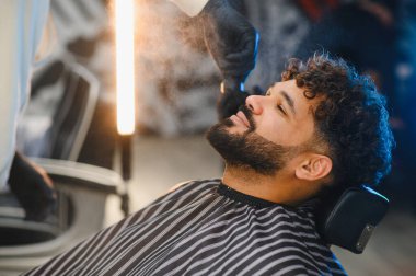 Berber dükkanında bakım hizmeti alan bir adam. Berber, kıvırcık saçlarına ve sakalına şekil vermesi için su püskürtüyor.