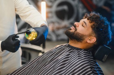 Berber bir adamın sakalına ve yüzüne rahatlatıcı sprey sıkar, berber dükkanında bakım ve bakım sağlar.