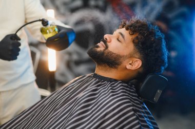 Sakallı, gözlerini kapayan bir adam berber koltuğunda dinlenirken berber yüzüne bir şekil ürünü püskürtüyor.