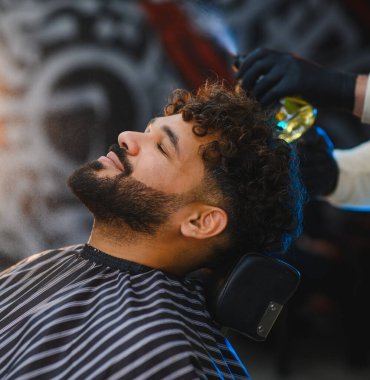 Gözleri kapalı bir adam kıvırcık saçlarını spreylerken profesyonel bir bakım deneyimi yaşıyor.