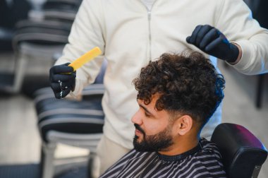 Berberin eldivenli elleri bir tarak ve makas tutuyor profesyonel bir berber dükkanında bir adamın kıvırcık saçlarını şekillendiriyor.