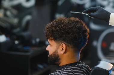 Modern bir berber dükkanında profesyonel saç kesimi hizmeti alan bir adam. Berberler makaslı, kıvırcık saçlı ve sakallı.