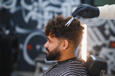 Kıvırcık saçlı ve sakallı bir adam berber dükkanında eldiven takarak berberden saç tıraşı oluyor.