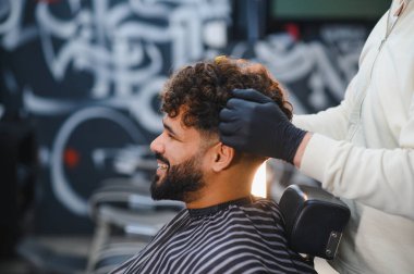 Gülümseyen adam profesyonel saç tıraşı oluyor ve siyah eldiven giyen bir berberden stil alıyor, tatmin oluyor.