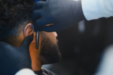 Siyah eldivenli berber bir adamın sakalını ve saçlarını geleneksel bir usturayla dikkatlice buduyor.