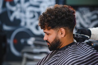 Kıvırcık saçlı ve sakallı bir adam berber dükkanında elektrikli makas kullanarak berberden yeni solmuş saç tıraşı oluyor.