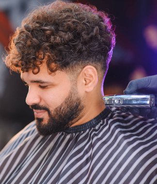 Kıvırcık saçlı ve sakallı berber müşterisi saç kesimi ve saç kesimi yaptırıyor.