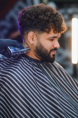 Berber, bir müşterinin sakalını ve yakasını elektrikli makasla buduyor, saçını yapıyor ve saçlarını tarıyor.