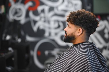 Sakallı ve kıvırcık saçlı bir adam berber dükkanında şık bir saç kesimi alıyor. Duvarın arkasında oturuyor.