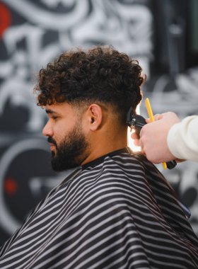 Kıvırcık saçlı ve sakallı berber müşterisi elektrikli makas kullanarak berberden yeni saç tıraşı oluyor.