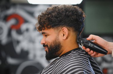 Berber, müşterinin kıvırcık saçlarını elektrikli makasla buduyor profesyonel bir berber dükkanında modern bir solgunluk stili yaratıyor.