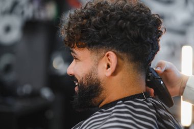 Profesyonel berber, berber dükkanında kıvırcık saçlarını ve sakallarını şekillendirmek için kırpıcı kullanırken mutlu adam gülümsüyor.