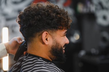 Müşteri, tıraş makinesiyle yeni saç kesiminin keyfini çıkarıyor. Profesyonel bir berber dükkanında kıvırcık bir bluz, soluk ve sakal gösteriyor.