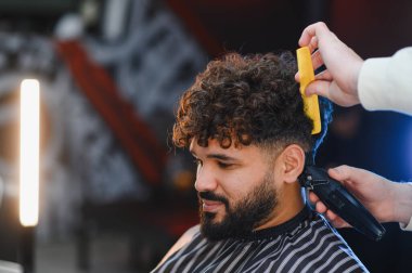 Berber, profesyonel bir berber dükkanında genç bir erkek müşterinin kıvırcık saçlarını taramak için tarak ve elektrikli makas kullanıyor.