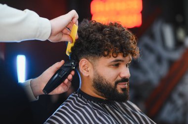 Kıvırcık saçlı bir adam berberden tıraş makinesi ve kuaförde tarak kullanıyor.