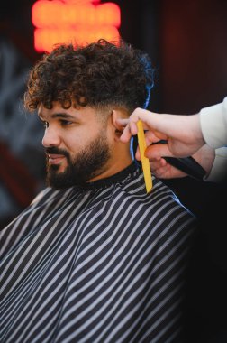 Kıvırcık saçlı ve sakallı bir adam profesyonel bir berber dükkanında tıraş makinesiyle yeni bir saç kesimi yaptırıyor.