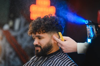 Kıvırcık saçlı bir adam yeni bir saç kesimi yaptırıyor, berber dükkanında stil yapıyor, saç yıkama spreyi kullanıyor.