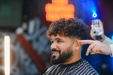 Gülümseyen adam, kuaför randevusu sırasında kıvırcık saçlarına profesyonel berber spreyi sıkar. Salon ortamında rahat ve kendinden emin görünür.