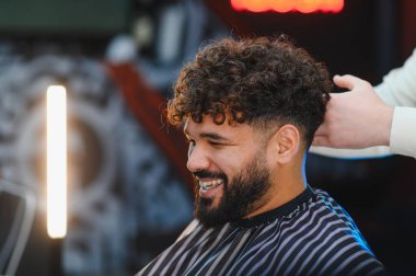 Berber saçını ve sakalını modern bir berber dükkanında şekillendirirken, mutlu adam gülümsüyor.
