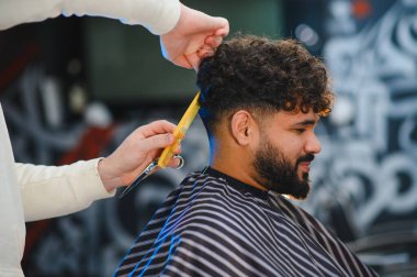 Bir adamın kıvırcık saçlarını ve sakalını makas ve tarak ile kesen berber stilisti, yeni bir görünüm sağlıyor.