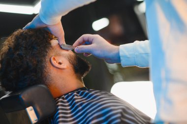 Profesyonel berber, modern bir berber dükkanında, bir müşterinin sakalını ustura ile dikkatlice traş ediyor, kuaförlük ve stil yapmaya odaklanıyor.