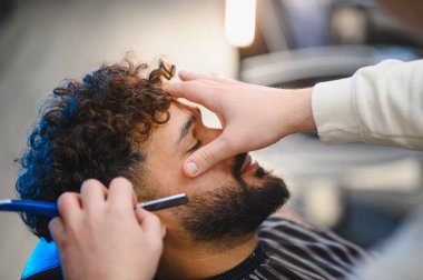 Kıvırcık saçlı bir adam berber dükkanında sakal bırakıyor. Berber traş olurken ustura kullanıyor.