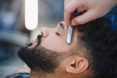 Profesyonel berber, berber dükkanında keskin bir usturayla bir adamın sakalını ve favorilerini dikkatlice şekillendiriyor.