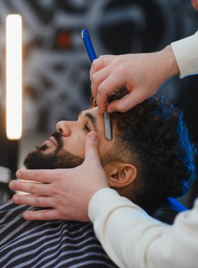 Berber, modern bir berber dükkanında erkek müşterinin sakal ve favorilerini keskin bir usturayla dikkatlice şekillendiriyor ve buduyor.