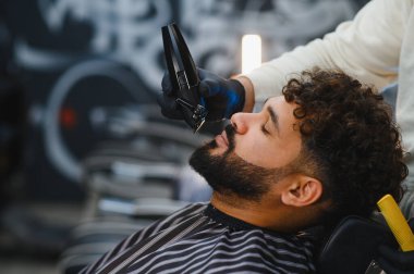 Müşteri berber dükkanında sakal tıraşı yaptırıyor. Berber, sakal ve saç stili için elektrikli tıraş makinesi kullanıyor.