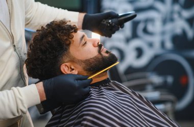 Berber müşterinin kıvırcık sakalını ve saçını kırpıcı ve tarak ile buduyor, profesyonel tımar ve stile odaklanıyor.