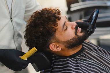 Berber, bir adamın sakalında elektrikli tıraş makinesi kullanıyor. Modern bir berber dükkanında kuaförlük ve kuaförlük hizmeti veriyor.