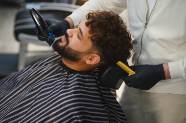 Berber, bir adamın sakalını tıraş etmek ve berber dükkanında kıvırcık saçlarını kesmek için elektrikli tıraş bıçağı kullanır.
