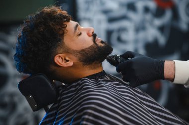 Profesyonel berber tıraş makinesi ve makasıyla bıyığını ve sakalını şekillendirirken kıvırcık saçlı adam dinleniyor.