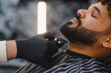 Berber, kuaförde adamın sakalını elektrikli jiletle buduyor. Temiz ve keskin bir görünüm için tımar ve tasarımı yakından takip ediyor.