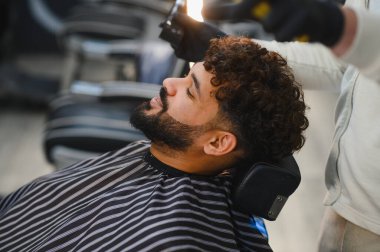 Profesyonel bir berber saç stili ve sakalı üzerinde çalışırken, kıvırcık saçlı bir adam berber koltuğunda dinleniyor.