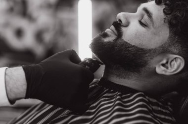 Berber, bir müşterinin sakalını elektrikli makasla dikkatlice buduyor ve modern bir berber dükkanında bakım hizmeti veriyor.