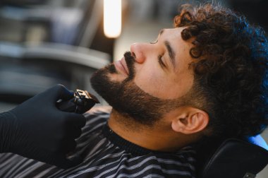 Berber koltuğunda dinlenen adam, profesyonel sakal kesme ve elektrikli makasla bakım hizmeti alıyor.