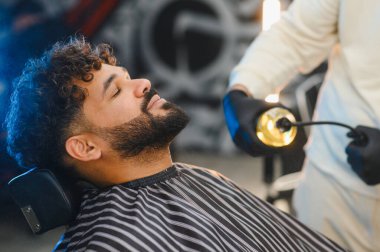 Kıvırcık saçlı ve sakallı bir adam berber dükkanında bakım hizmeti alıyor, berber sakalına şişe sıkıyor.